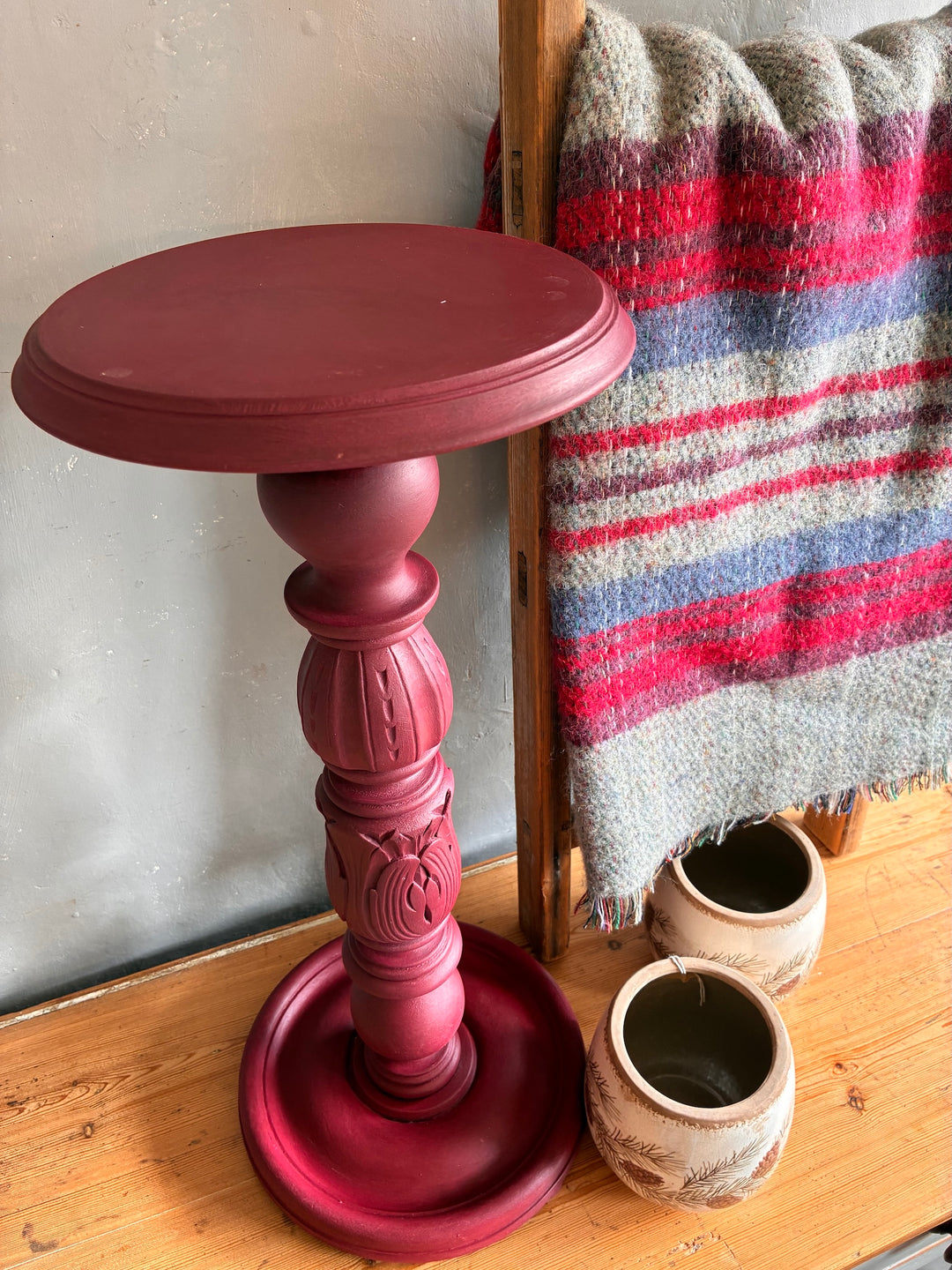 Vintage Wooden Burgundy Plant Stand with red top and beautifully carved base next to woven blanket and pots.