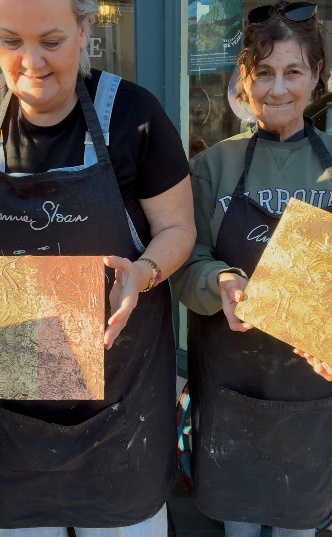 Two Annie Sloan chalk paint workshop attendees wearing aprons holding wooden boards with gold leaf designs at Source for the Goose, Devon