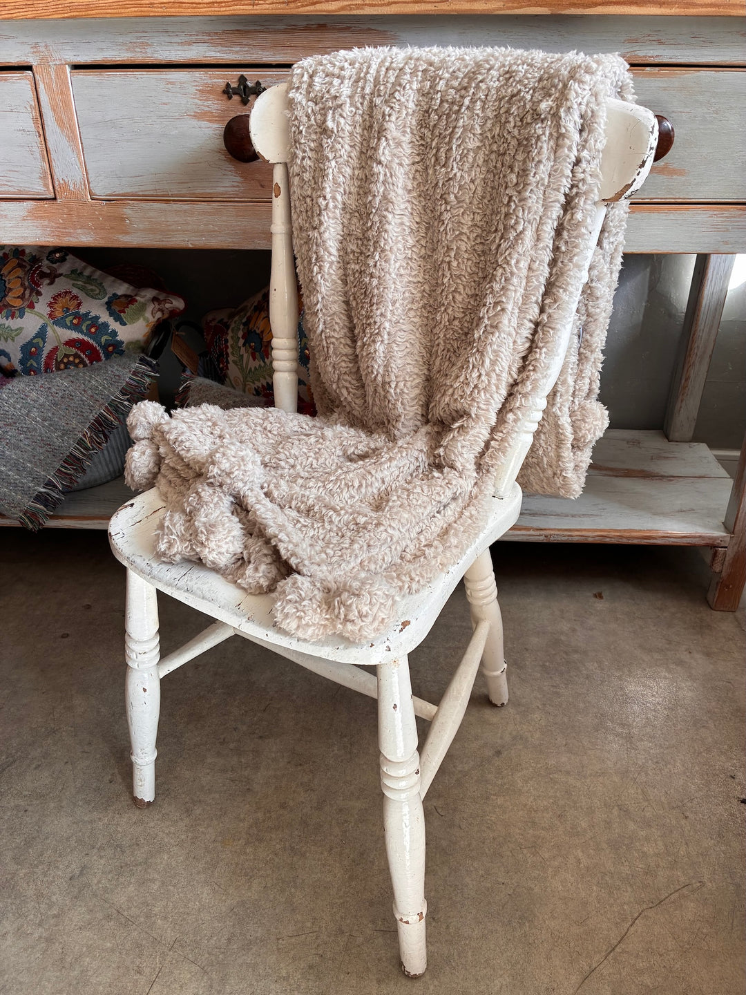 Cosy Pom Pom Throw in Linen draped over a white chair, showcasing its soft texture and charming pom poms.