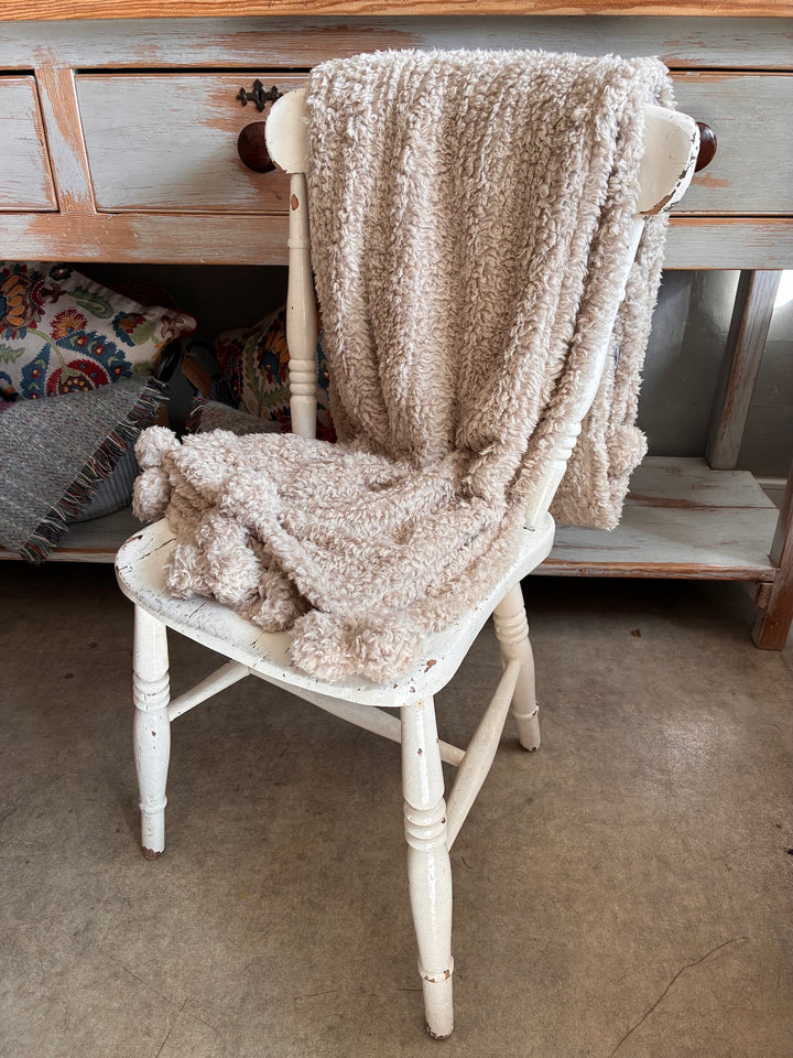 Cosy Pom Pom Throw in Linen draped over a white chair, showcasing its soft texture and charming pom poms.