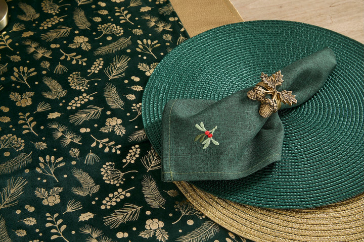 A dark green woven placemat with a gold botanical pattern, styled with a matching green napkin featuring a small embroidered bird and berry motif, and a round woven straw placemat underneath.
