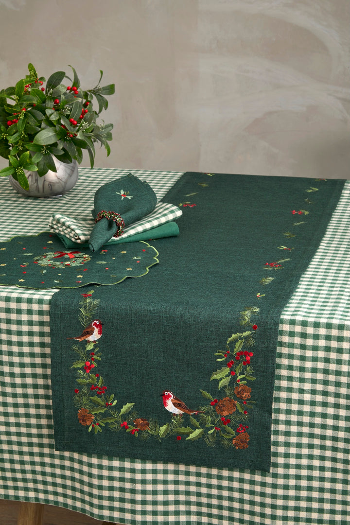 A dark green table runner with embroidered birds, holly leaves, and red berries, styled on a green and white checkered tablecloth with matching napkins and a potted plant.