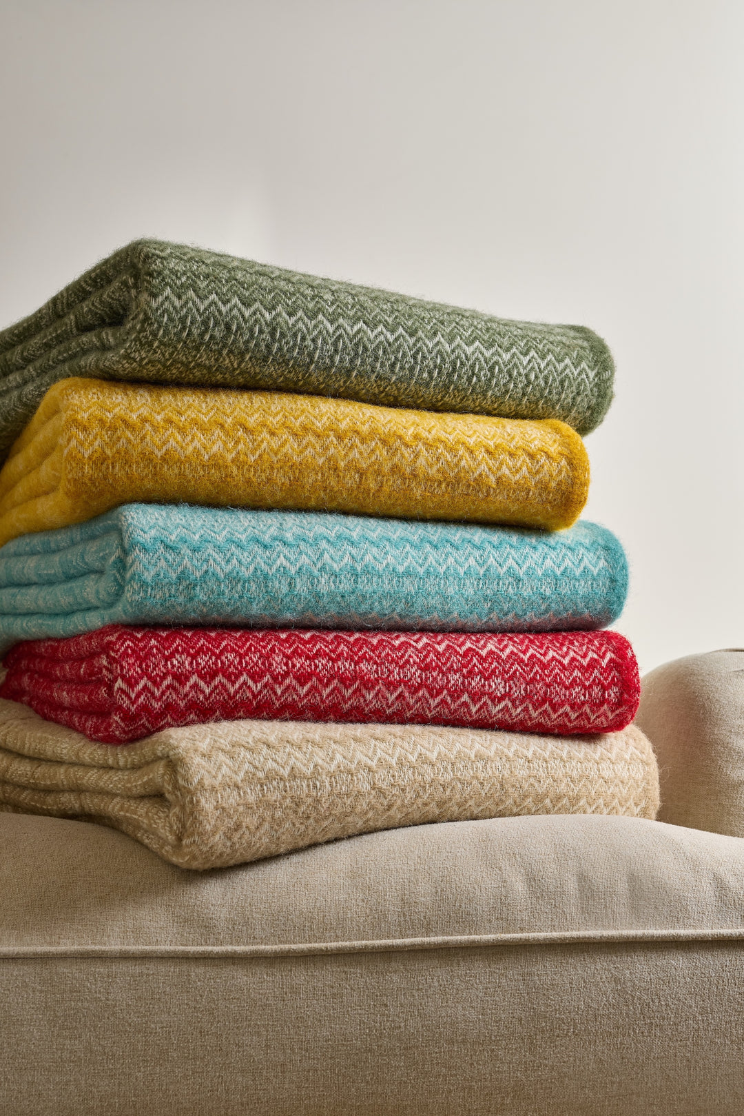 Stack of colorful blankets including the Tweedmill Oatmeal Fair Isle Wool Throw on a sofa.