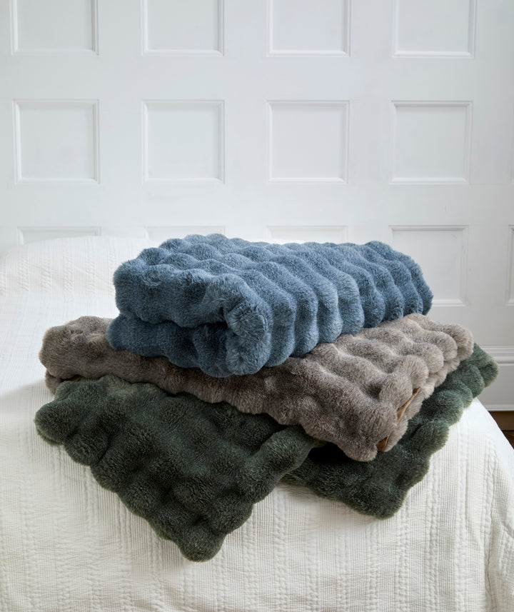 Slate Blue Faux Fur Throw draped among other luxurious throws on a bed, showcasing its soft texture and cozy appeal.