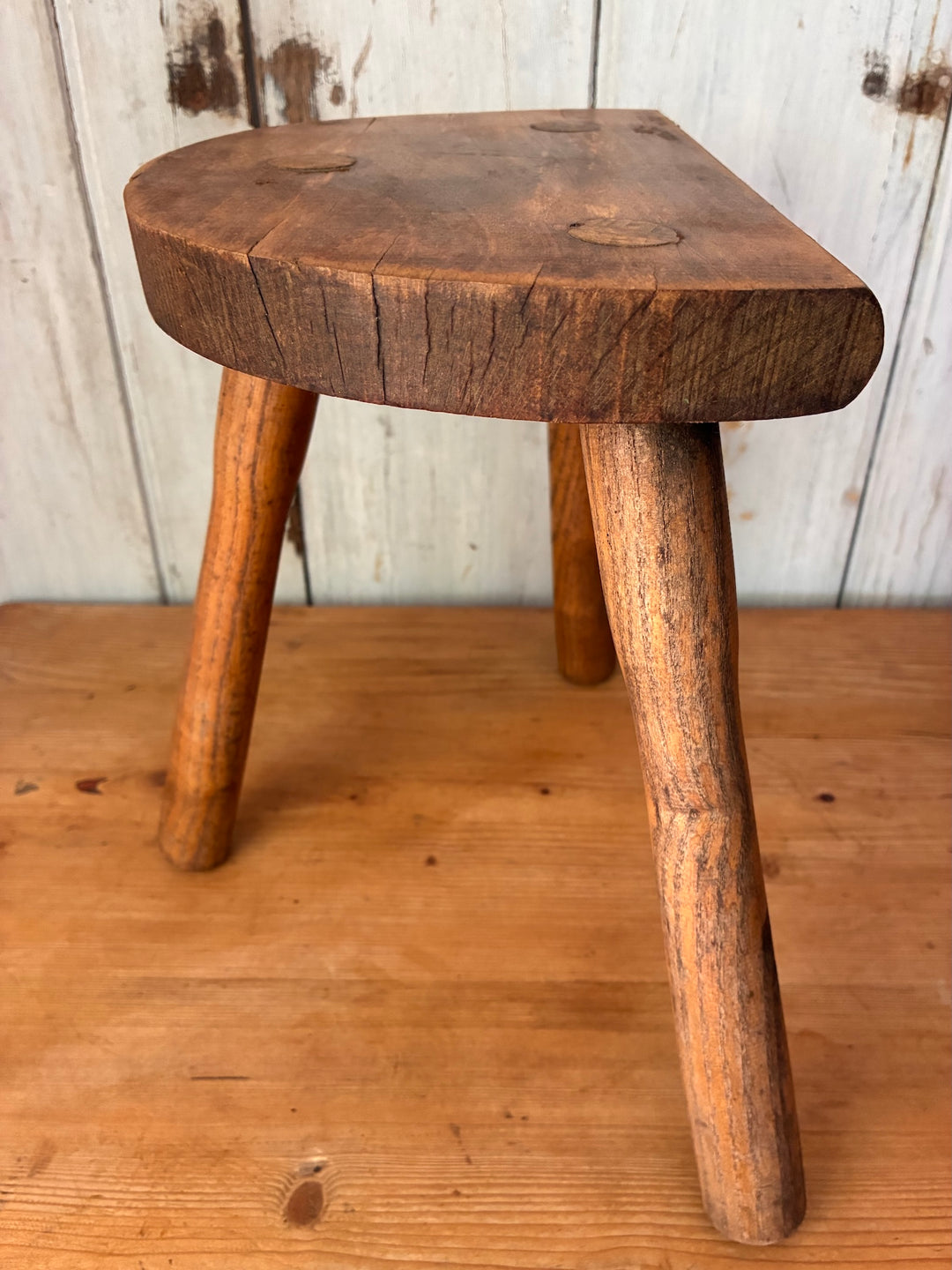 Vintage Wooden French Tripod Stool with rustic wooden legs and demi-lune seat on a wooden background.