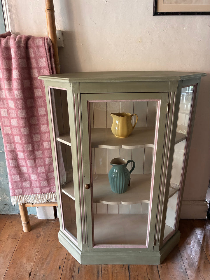 Soft Green Corner Display Cabinet with glass doors and pink accents, featuring colorful pitchers inside. Source for the Goose, Devon.