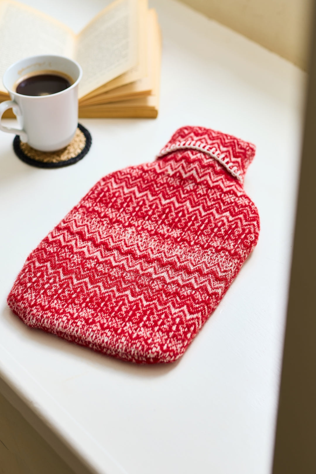 Fair Isle Wool Hot Water Bottle - Two Colours on a table with a coffee mug and a book, showcasing heritage style for a cosy home.