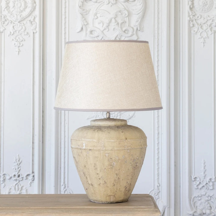 Marius Lamp - Stoneware Base With Woven Linen Shade on a wooden table against a textured white backdrop.