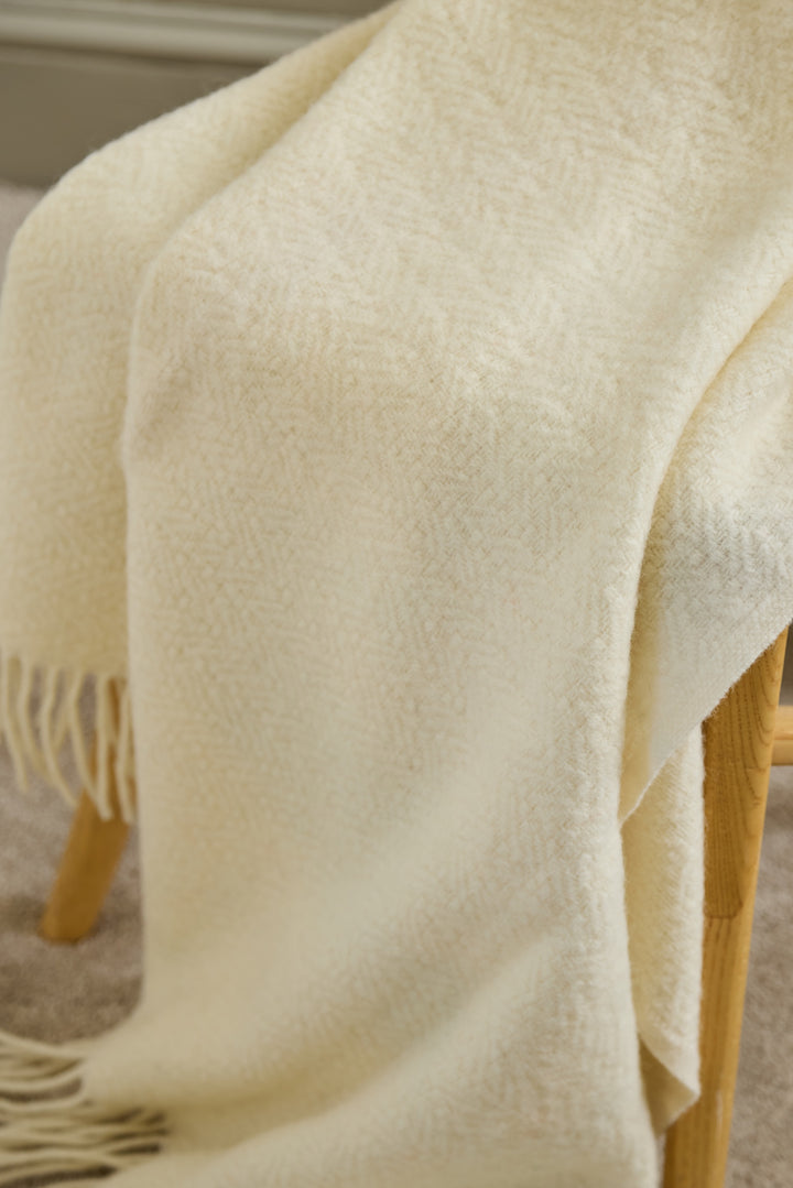 The Moorland - Cream Wool Throw draped over a chair, showcasing its soft texture and herringbone pattern in a cosy home.