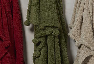 Cosy Pom Pom Throw in Olive Green showcasing plush fabric and playful pom poms, hung among other colors.