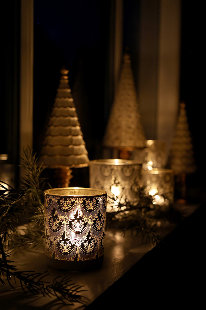 Pretty glass hurricane candle holder with white lace design, reflecting warm candlelight near decorative Christmas trees.