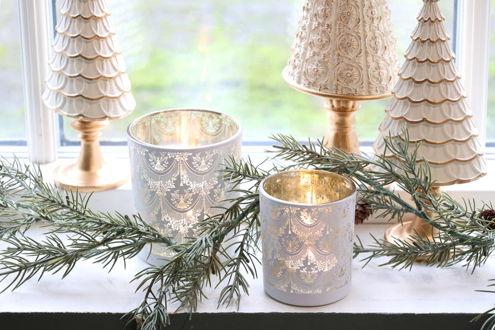 Pretty glass hurricane candle holders with lace designs, surrounded by greenery and decorative trees.