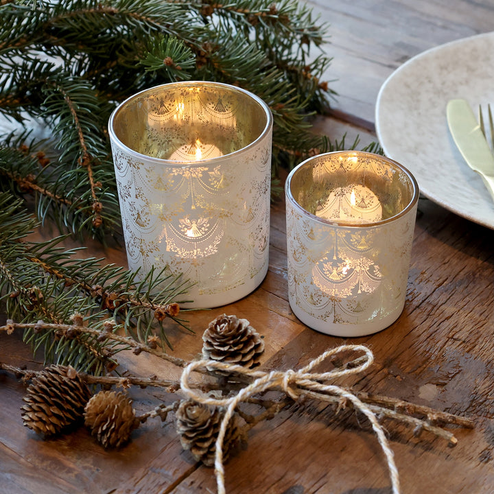 Pretty glass hurricane candle holders with white lace design, glowing warmly on a wooden table with pinecones and greenery.