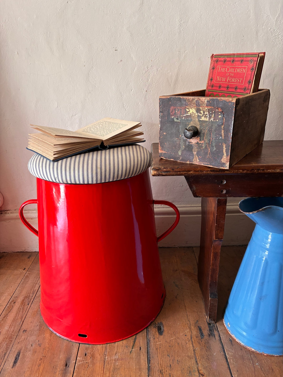 Quirky red enamel stool with blue stripe ticking lid and an open book, perfect for stylish storage in your home.