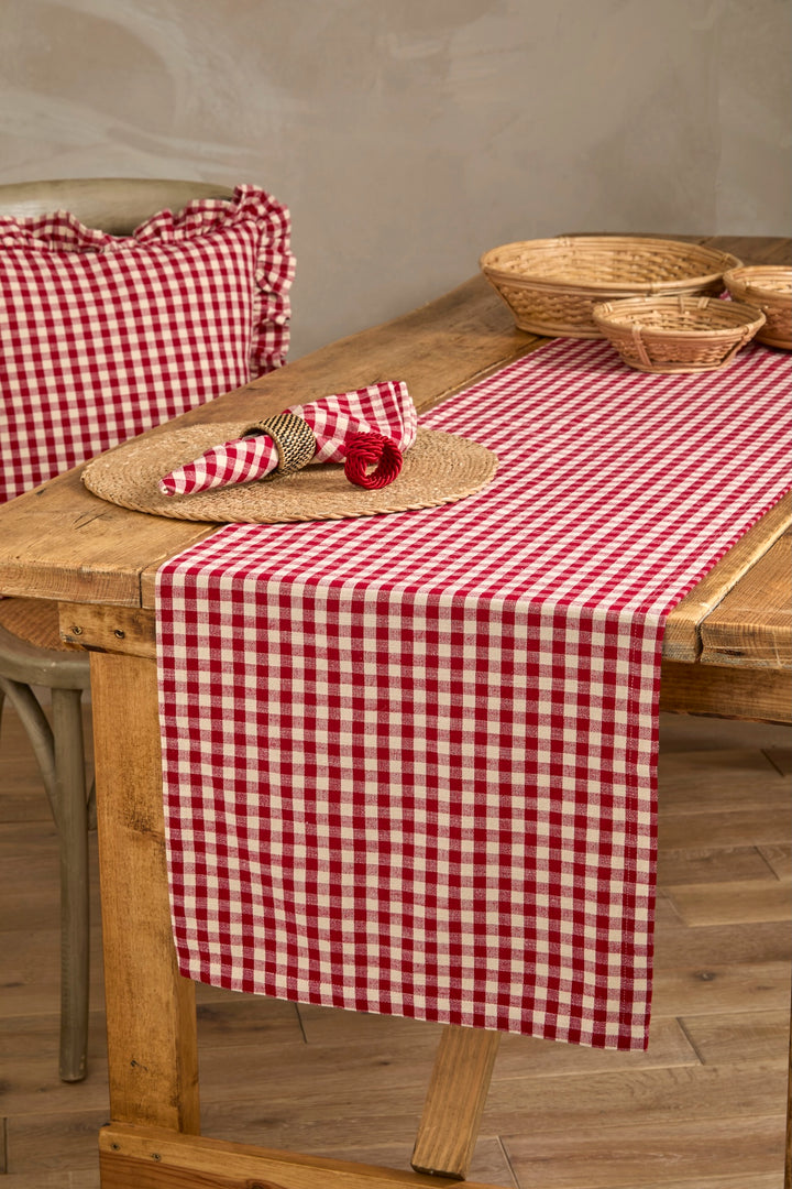 A red and white gingham check table runner draped over a rustic wooden dining table, styled with a matching pillow, woven placemat, napkin, and small red flower, with baskets in the background.