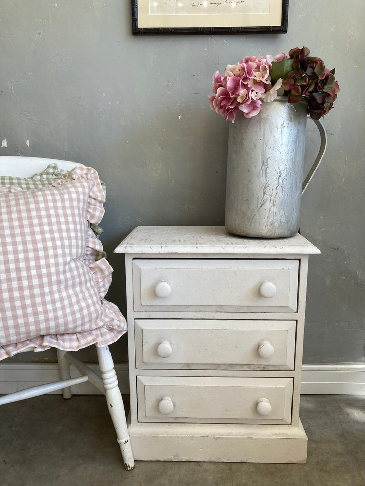 Charming white bedside table with three drawers, pink flowers in a metal jug, and a cozy checkered pillow.