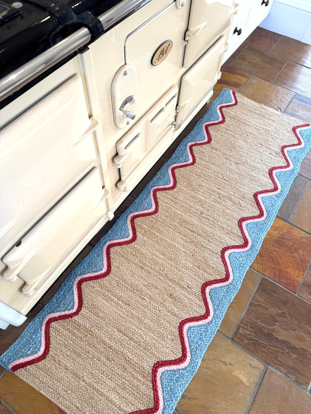 Sky Wave Organic Jute Braided Rug placed in front of a stove, showcasing its sky blue wave border and soft pink trim.