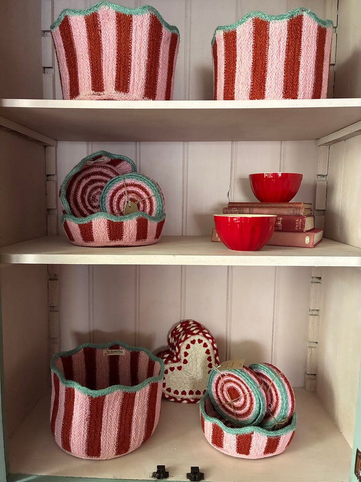 A set of handmade woven storage baskets and bowls in pink, red, and mint green, displayed on white shelves against a white paneled wall.
