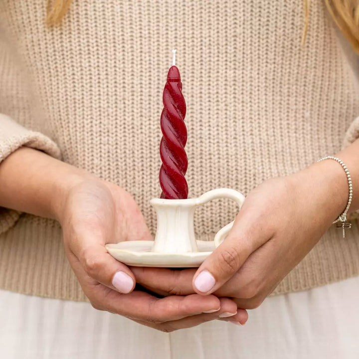 Woman holding a twisted plum red dinner candle in a cream-colored holder, showcasing sophisticated table decor. Source for the Goose Devon.