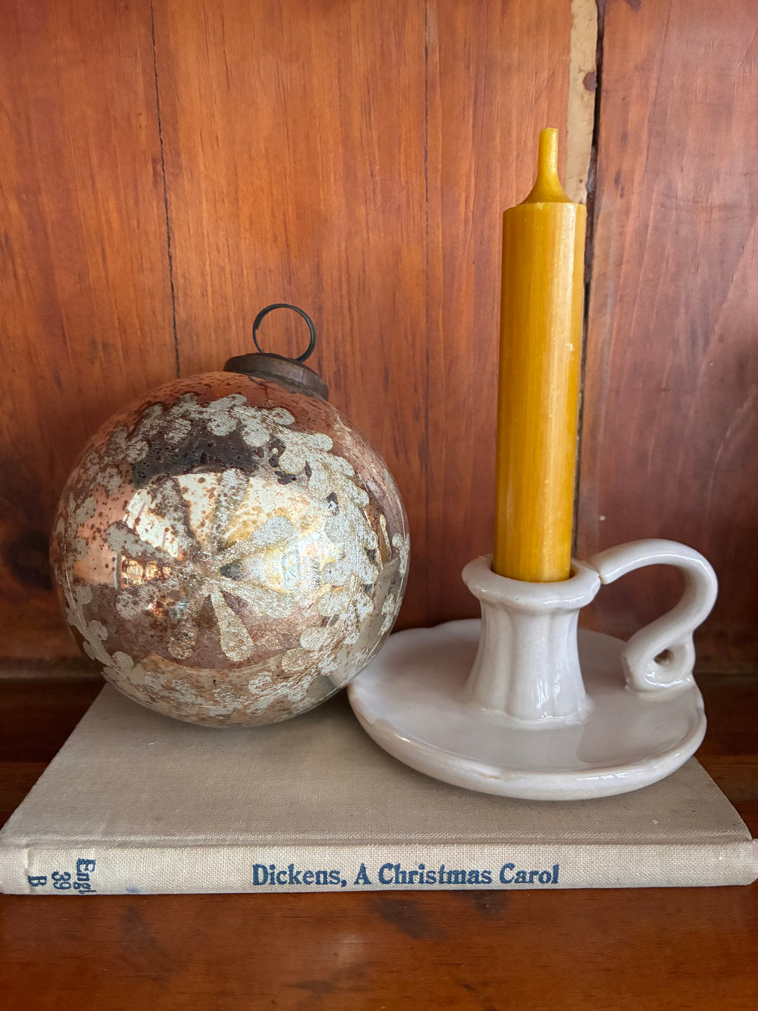 Vintage Style Wreath Design Glass Bauble beside a white candle holder on a book of Dickens, A Christmas Carol.