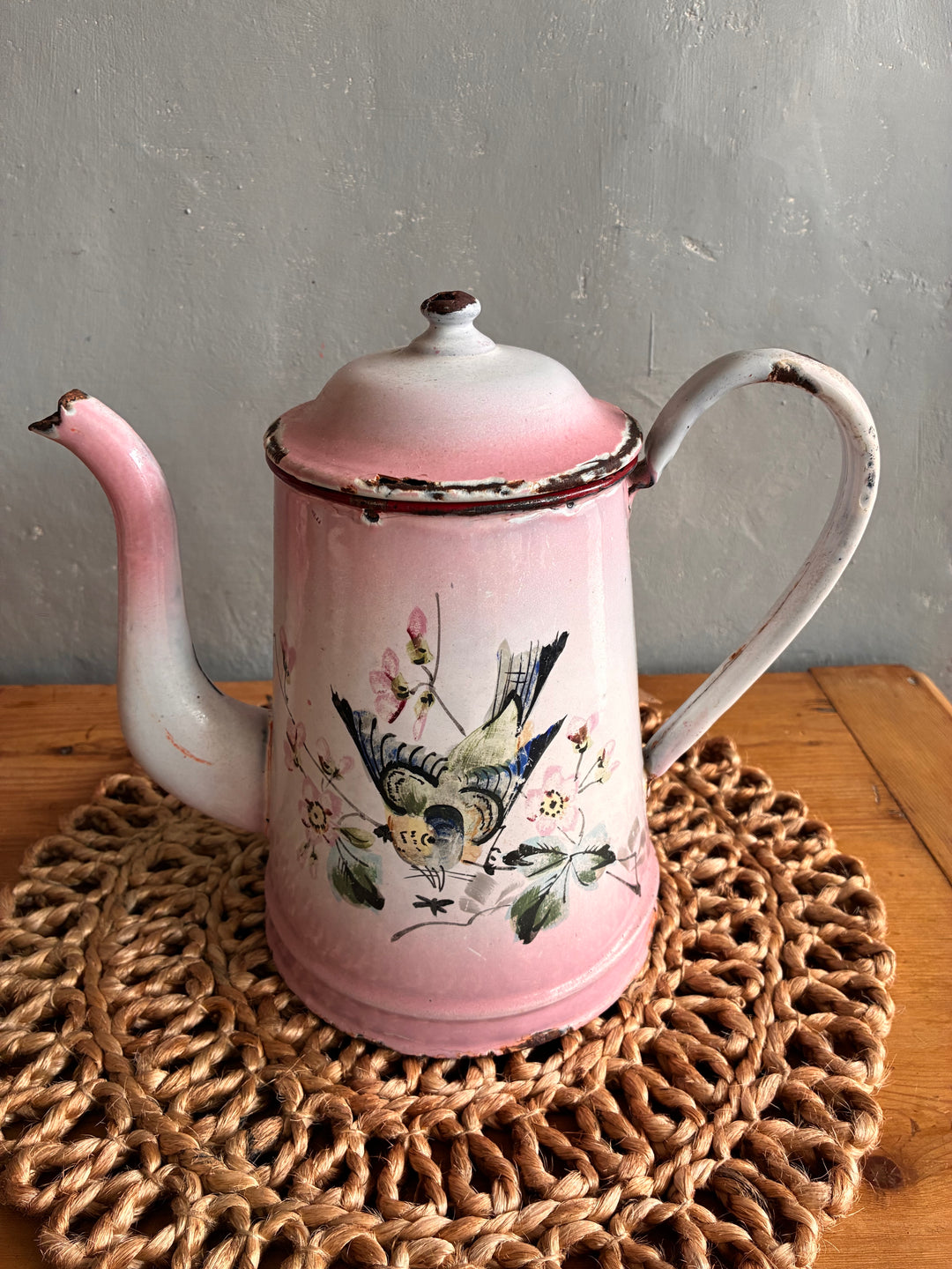 Vintage pink and white French enamel coffee pot with hand-painted bird design on a woven placemat.