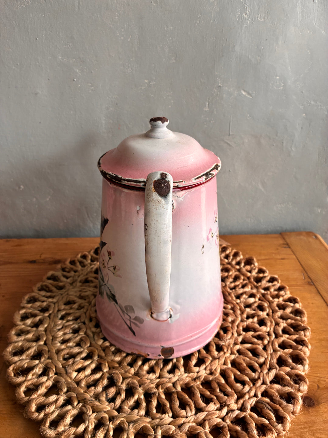 Vintage pink and white French enamel coffee pot with hand-painted bird design on a woven placemat.