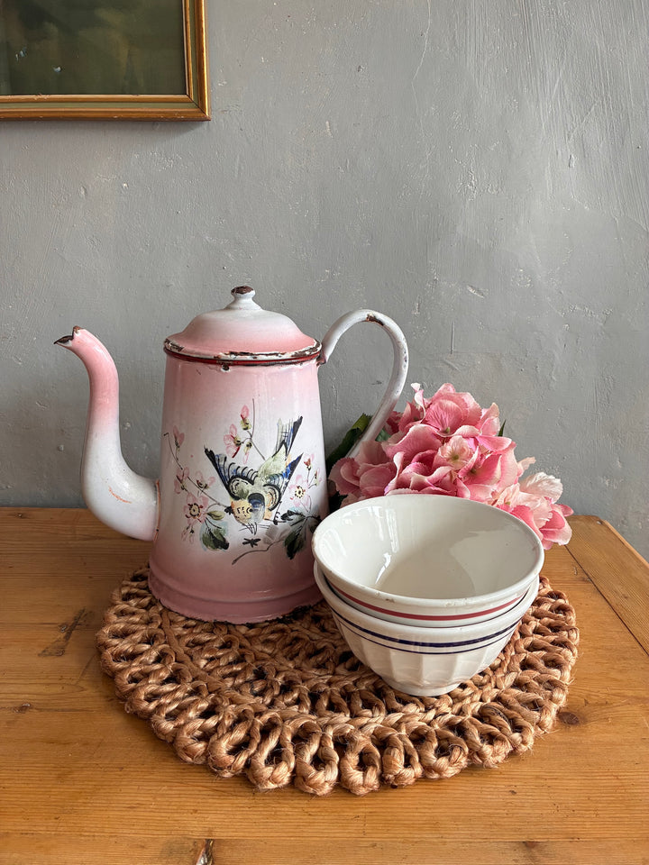 Vintage pink and white French enamel coffee pot with hand-painted birds beside a bowl and flowers on a woven placemat.