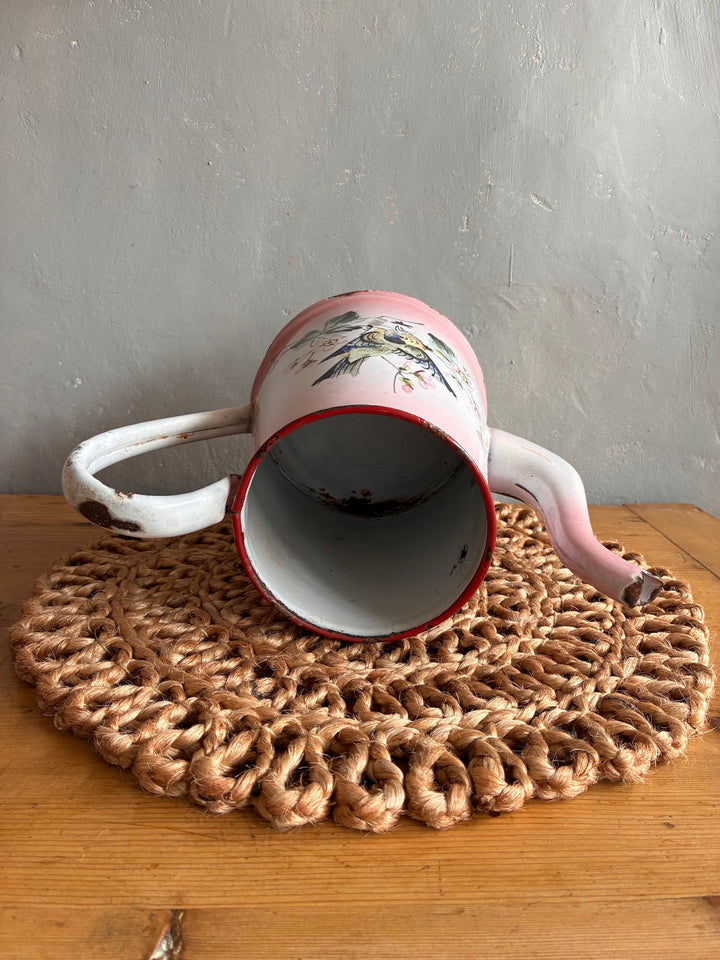 Vintage pink and white French enamel coffee pot with bird design on a woven mat, perfect for nostalgic kitchen decor.