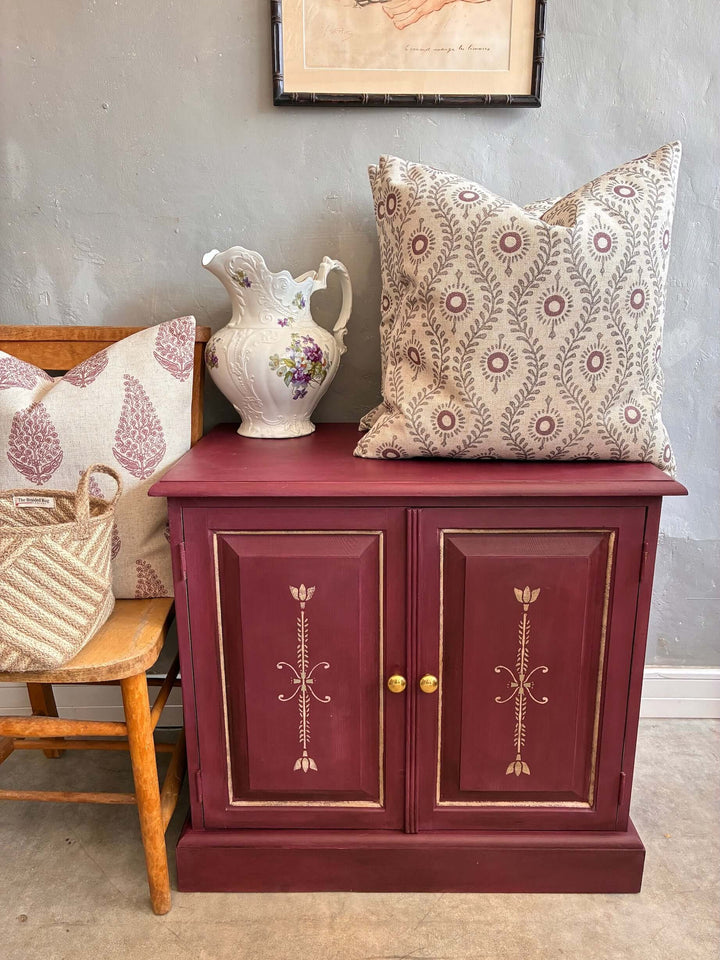 Aubergine folk art style cupboard with decorative pillow and pitcher, perfect for stylish small space storage.