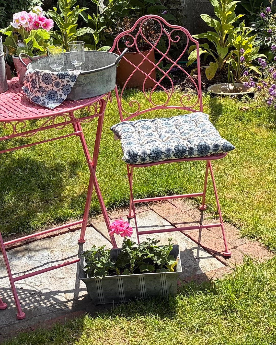 Zinc Ornate Planter With Feet alongside a pink chair and table in a vibrant garden setting.