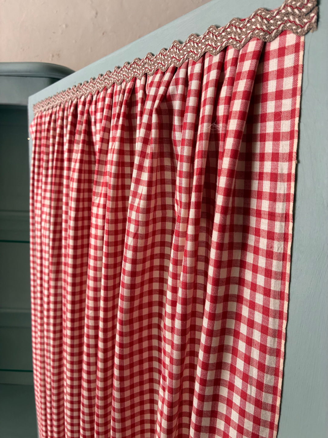 Close-up of red gingham curtain with decorative trim on a Small Blue Painted French Armoire by Source for the Goose, Devon.