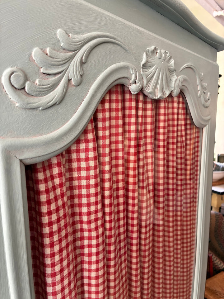 Close-up of the Small Blue Painted French Armoire showcasing the ornate top and red gingham curtain.