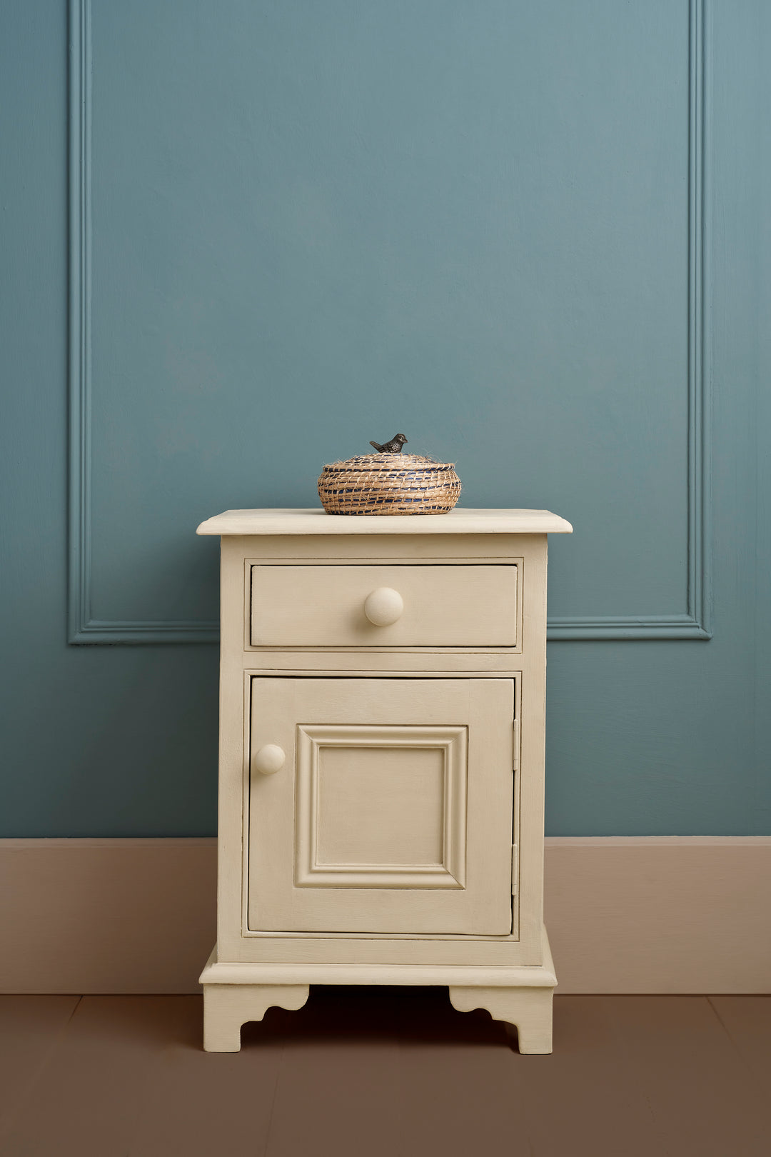 Elegant putty-toned bedside table against a blue wall, showcasing a rustic decorative basket on top.