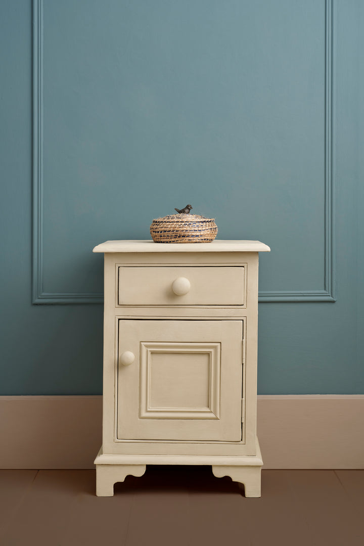 Elegant putty-toned bedside table against a blue wall, showcasing a rustic decorative basket on top.