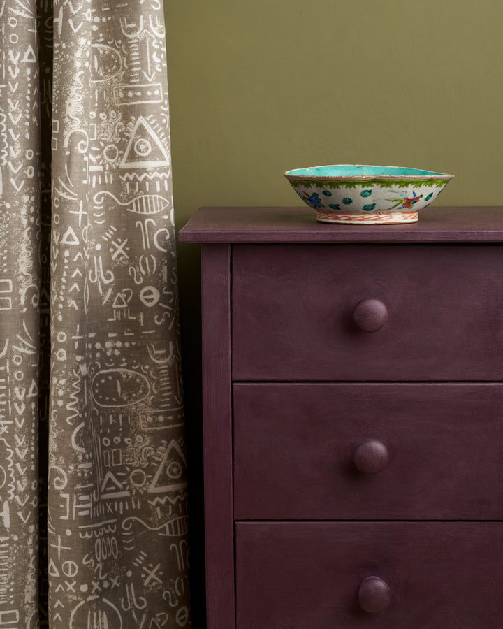 Tyrian Plum Chalk Paint® applied to a bedside cabinet, paired with patterned curtains and decorative bowl.