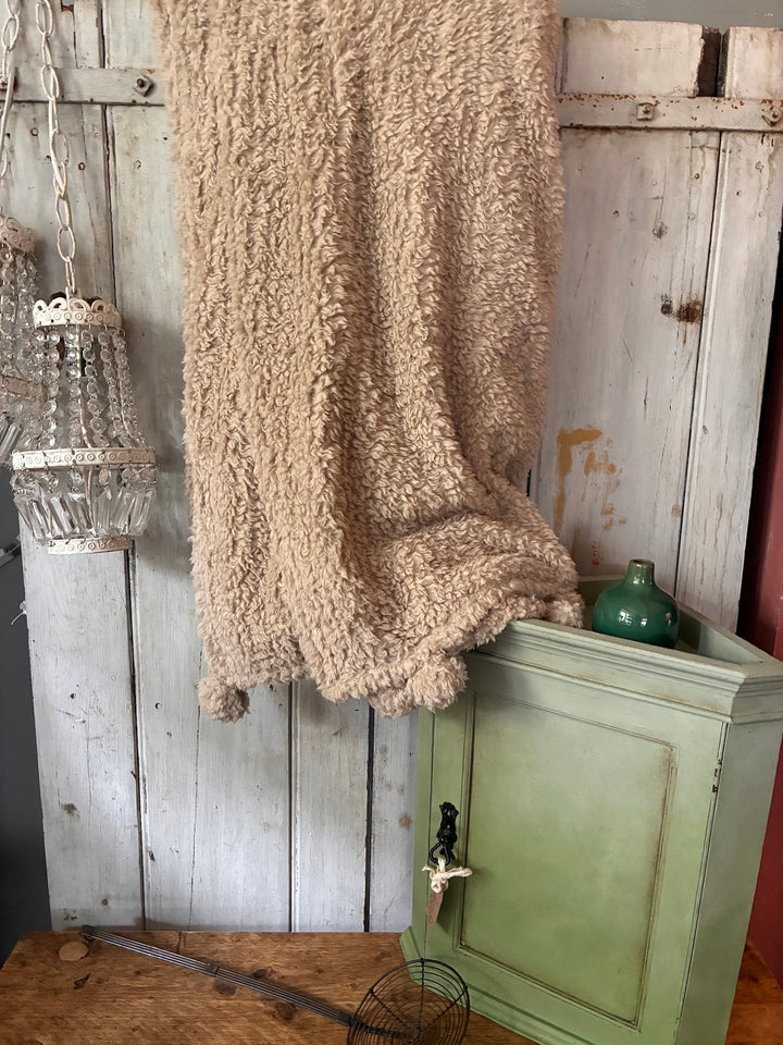 cosy throw with pom pom ends in a soft linen colour against a wooden wall with a green cupboard in front