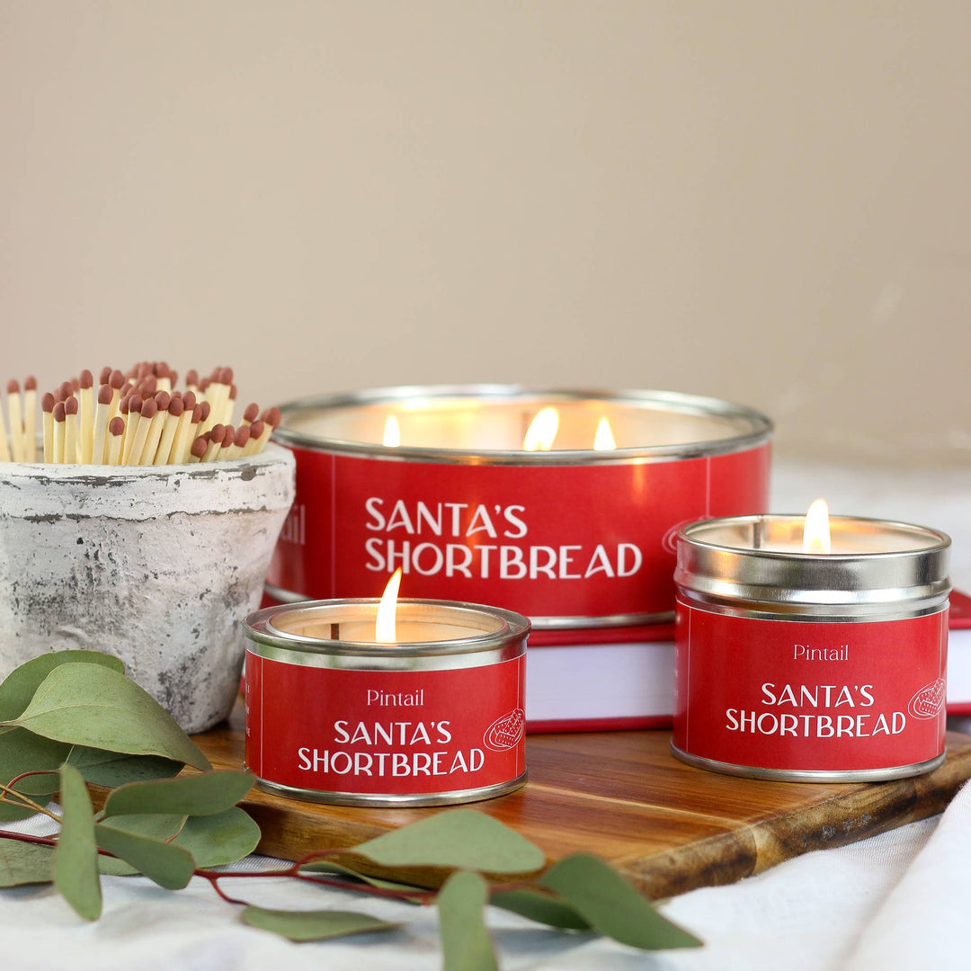 Santa's Shortbread Classic Tin Candle by Pintail Candles, lit and displayed with matches, offering a cosy festive atmosphere.