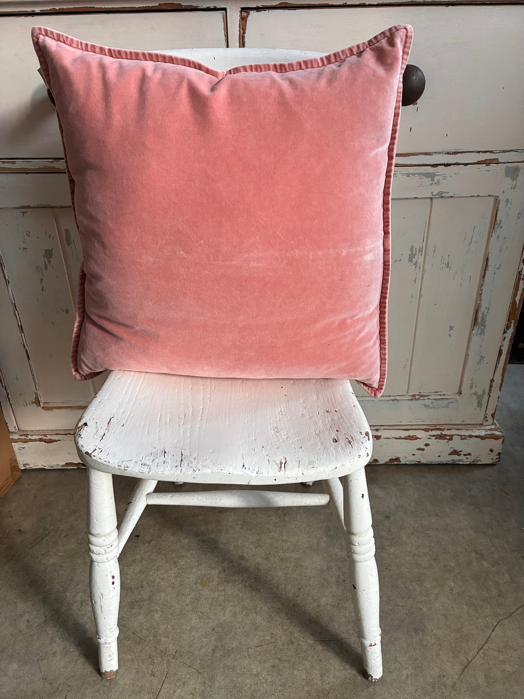 Pink cushion on a white distressed chair against a wooden wall.