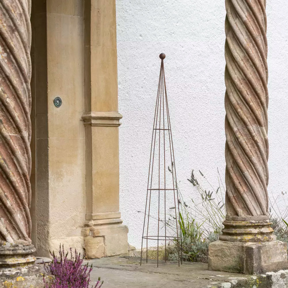 Metal garden obelisk plant support with antique brown finish, placed by entrance, enhancing outdoor aesthetics.
