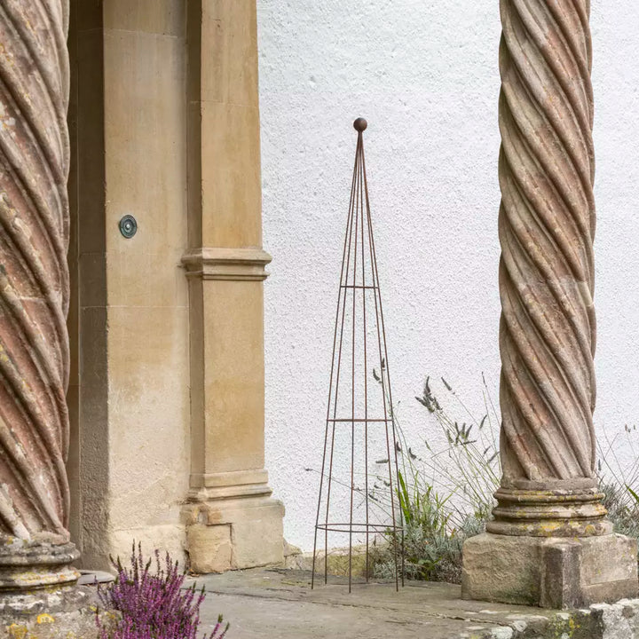 Metal garden obelisk plant support with antique brown finish, placed by entrance, enhancing outdoor aesthetics.