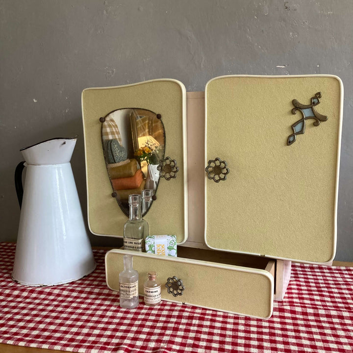 Mid Century French wall cabinet with a pear-shaped mirror, filigree handles, and pale yellow veneer on a checked tablecloth. Source for the Goose Devon.
