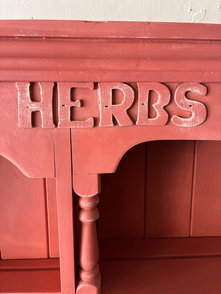 Close-up of the raised 'HERBS' lettering on a salmon-toned pink wall-mounted decorative rack.