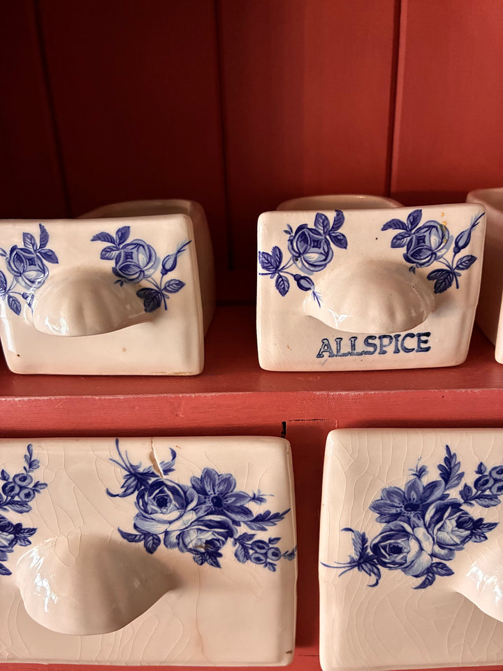 Ceramic spice drawers with blue floral designs, featuring 'Allspice' label, on a rustic wall-mounted rack.
