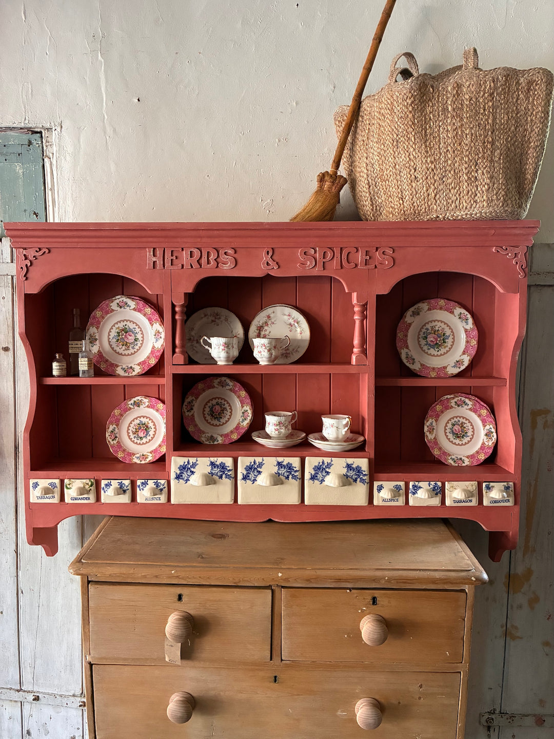 Wall-mounted plate rack in salmon pink featuring herbs & spices drawers, displaying elegant dishware and ceramic spice containers.