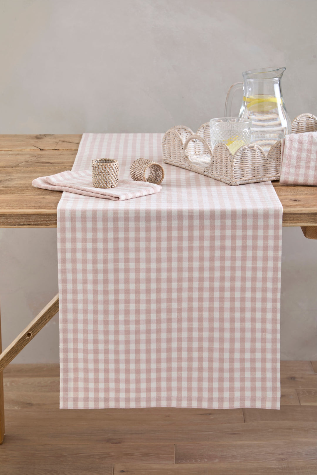 Plaster Pink Gingham Table Runner elegantly displayed on a wooden table with glassware and woven accessories.