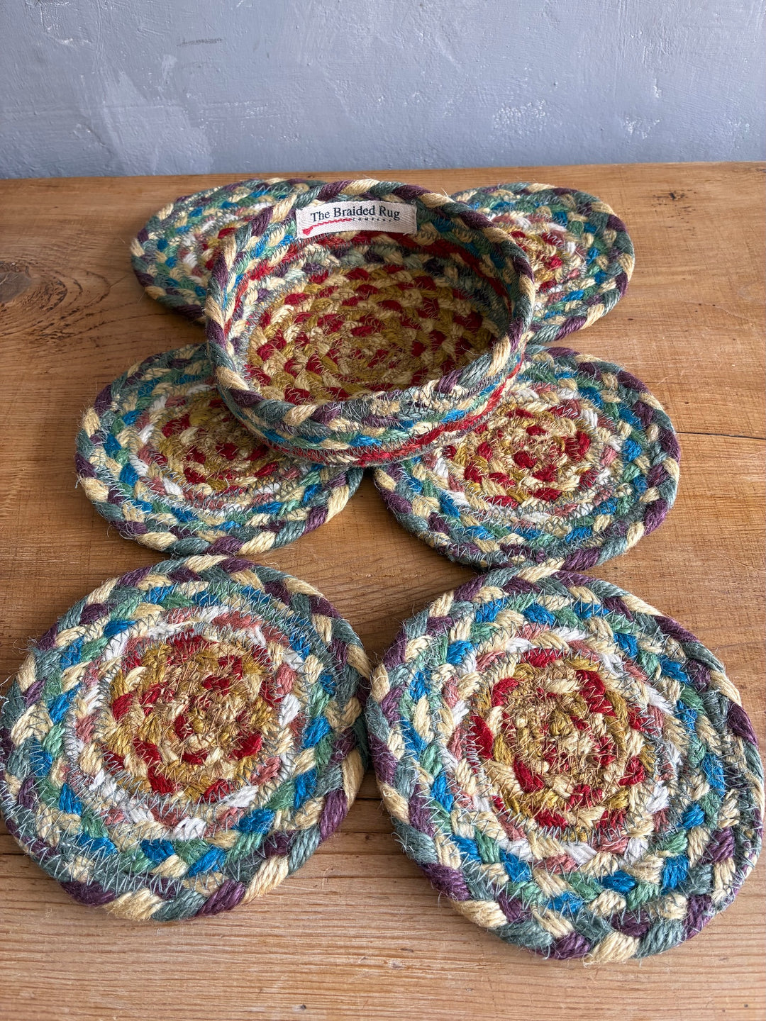 Five round braided jute coasters in a multicolor pattern of blue, red, beige, and purple, arranged on a wooden tabletop with a gray wall behind them.