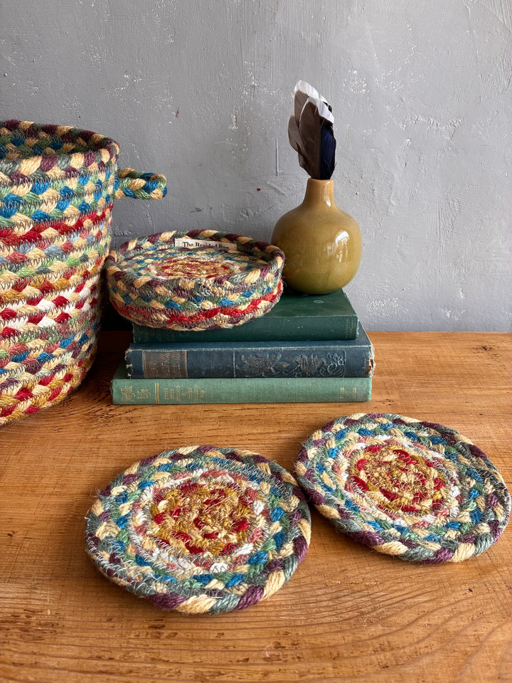 A set of handmade braided jute coasters in a multicolor pattern of blue, red, yellow, and green, displayed on a wooden table with books and a ceramic vase in the background.