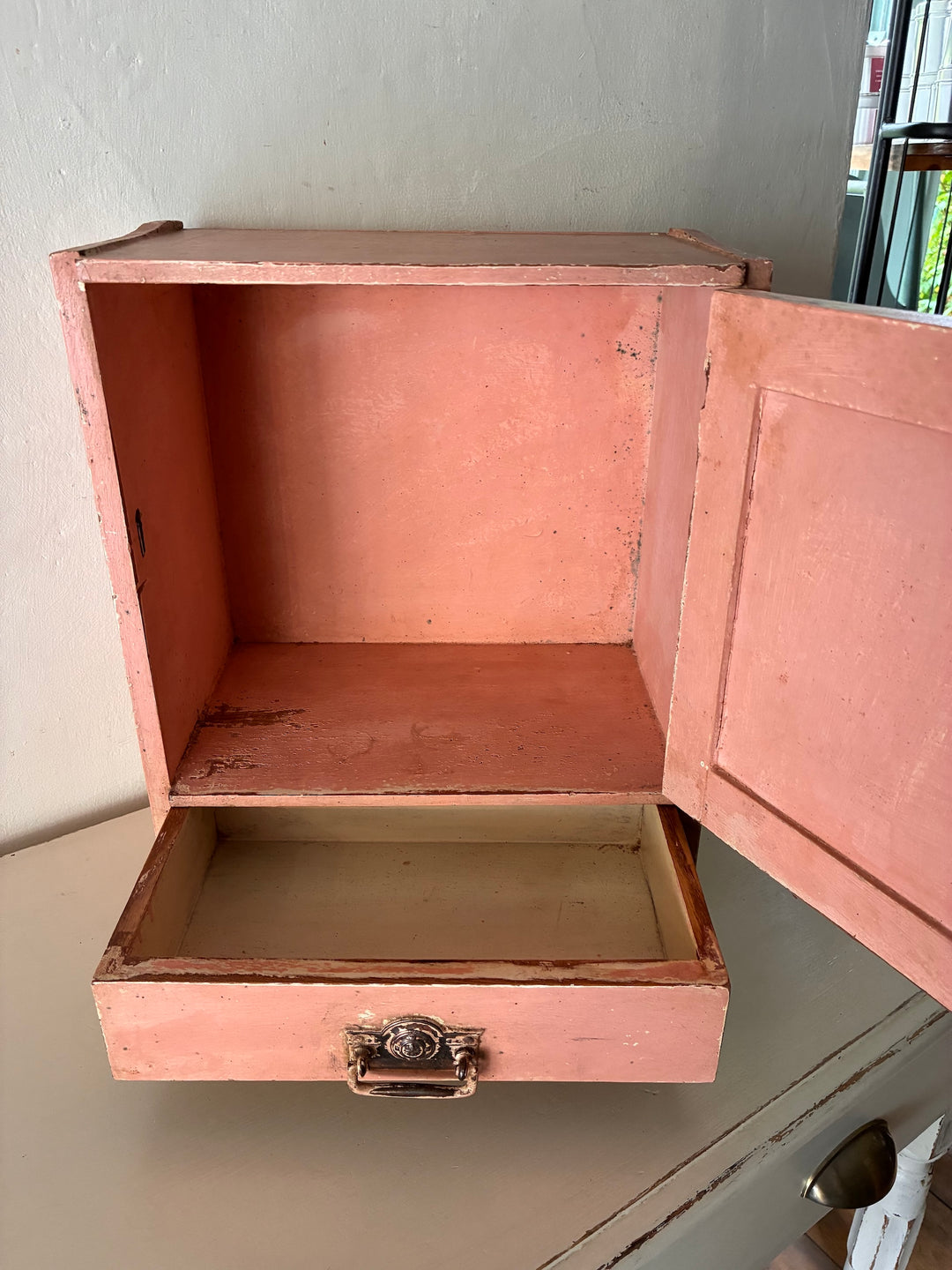 Small vintage salmon pink wall cupboard with distressed finish and brass handles, perfect for rustic kitchens and bathrooms.