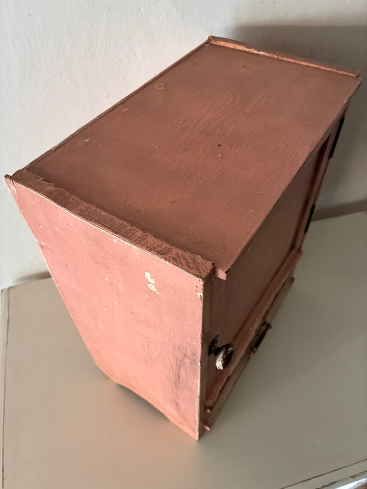 Top view of a small vintage salmon pink wall cupboard with distressed paint and a brass handle, by Source for the Goose, Devon.