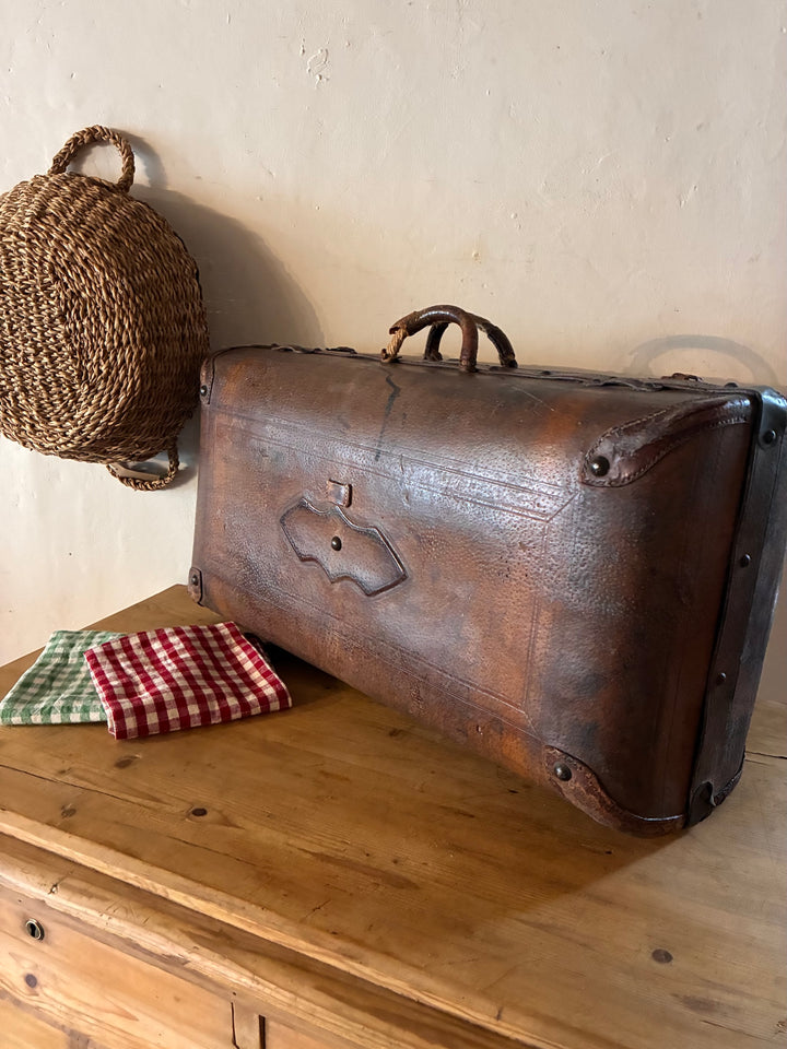 Vintage French Leather Suitcase resting on a wooden surface with a woven basket and checkered cloths in the background.